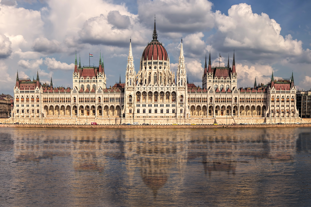 Hungarian Parliament Building along the Danube with a blue sky and fluffy clouds, reflected in the water below.