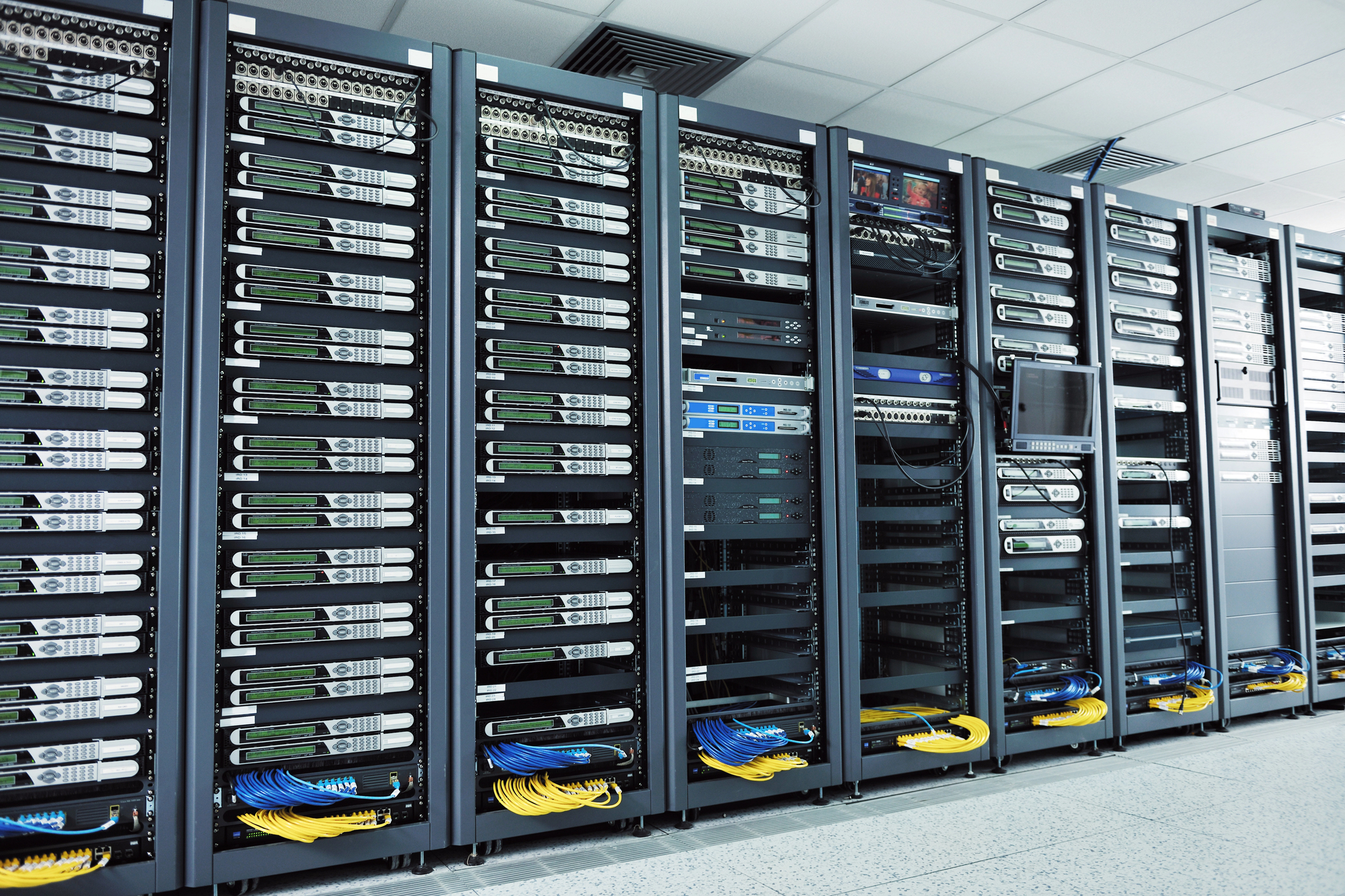 Row of rack-mounted servers in a data center with blue and yellow network cables at the base and green LCDs on the front panels.