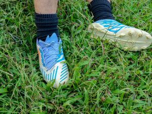 Blue and white soccer cleats on feet standing in tall green grass (worn with dark socks).