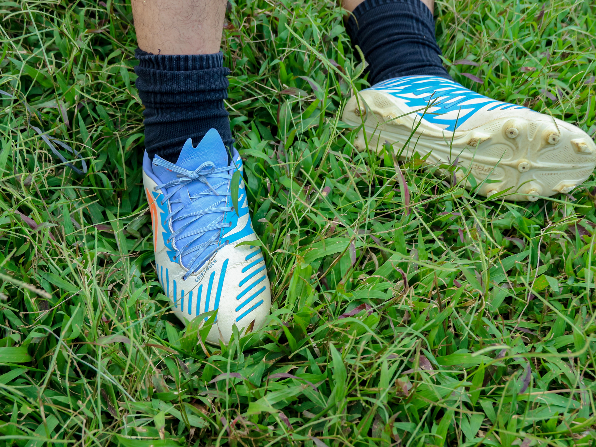Blue and white soccer cleats on feet standing in tall green grass (worn with dark socks).