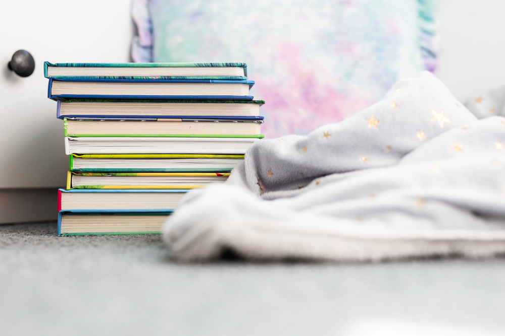 Stack of colorful hardcover books stacked on a carpeted floor beside a soft gray blanket with gold stars at the edge of the frame.