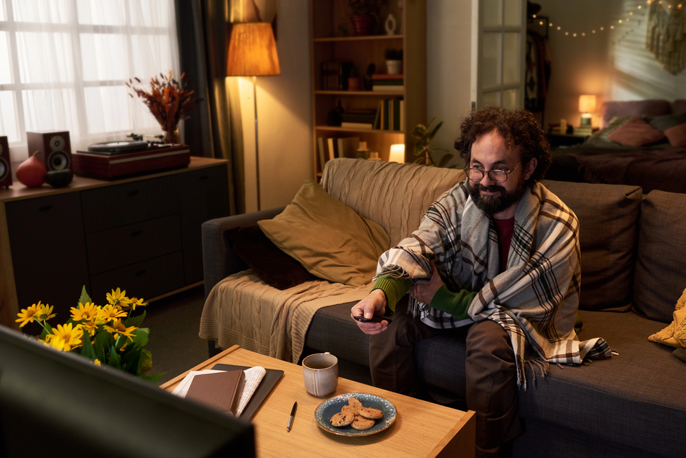 Bearded man in a plaid blanket sits on a sofa, remote in hand, watching TV with cookies and a mug on the table nearby.