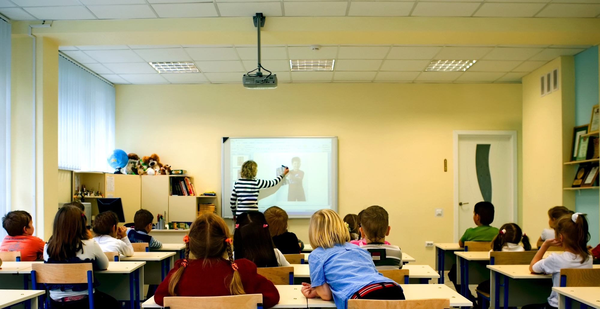 Students seated at desks in a classroom, a teacher writes on an interactive whiteboard at the front.