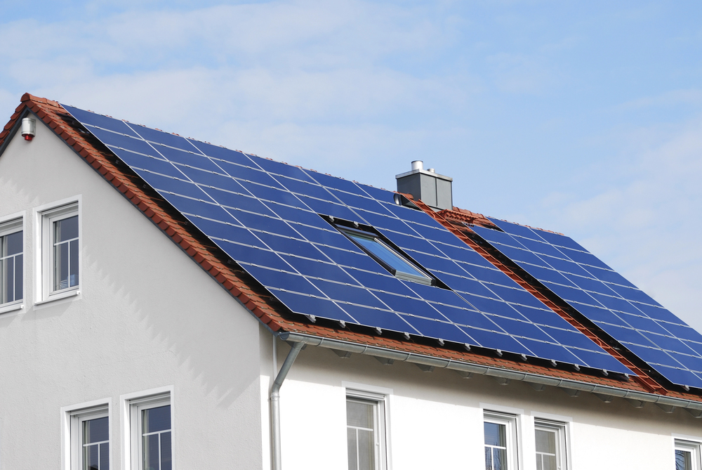 White two-story house with dark blue solar panels covering the roof under a blue sky.
