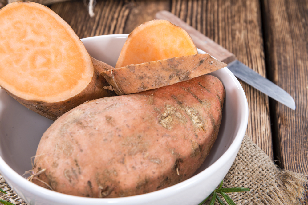 White bowl containing a whole and sliced orange-skinned sweet potato on a rustic wooden table, ready for cooking.