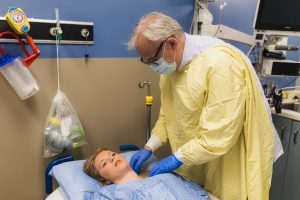 Hospital scene: clinician in a yellow gown and mask tending to a patient lying in bed.