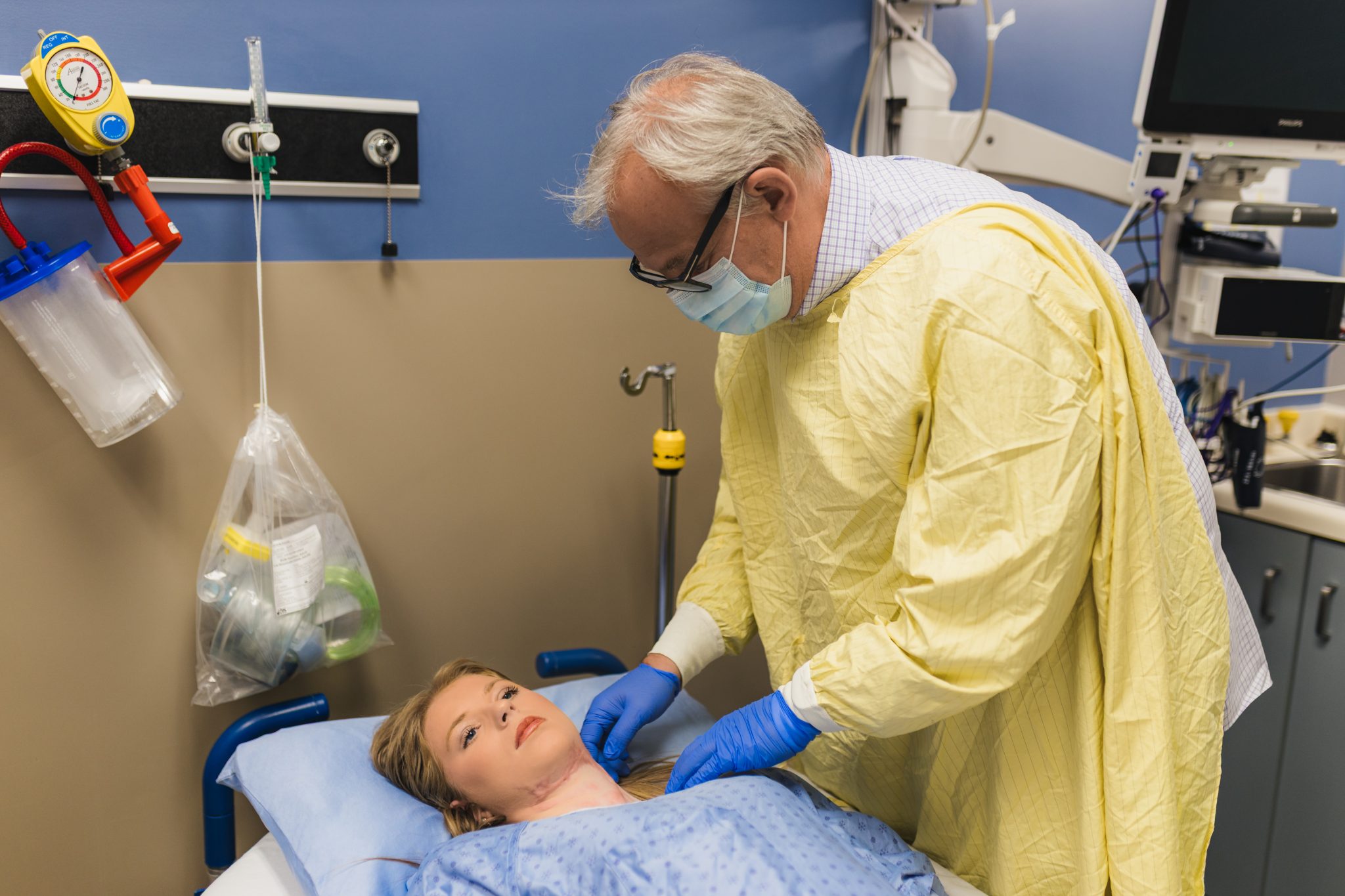 Hospital scene: clinician in a yellow gown and mask tending to a patient lying in bed.