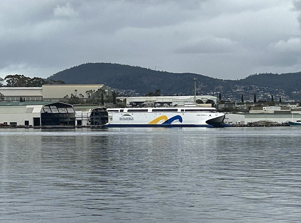 White passenger ferry docked at a marina, featuring blue and yellow wave graphics on its side, with calm water and industrial buildings in the background and hills beyond.