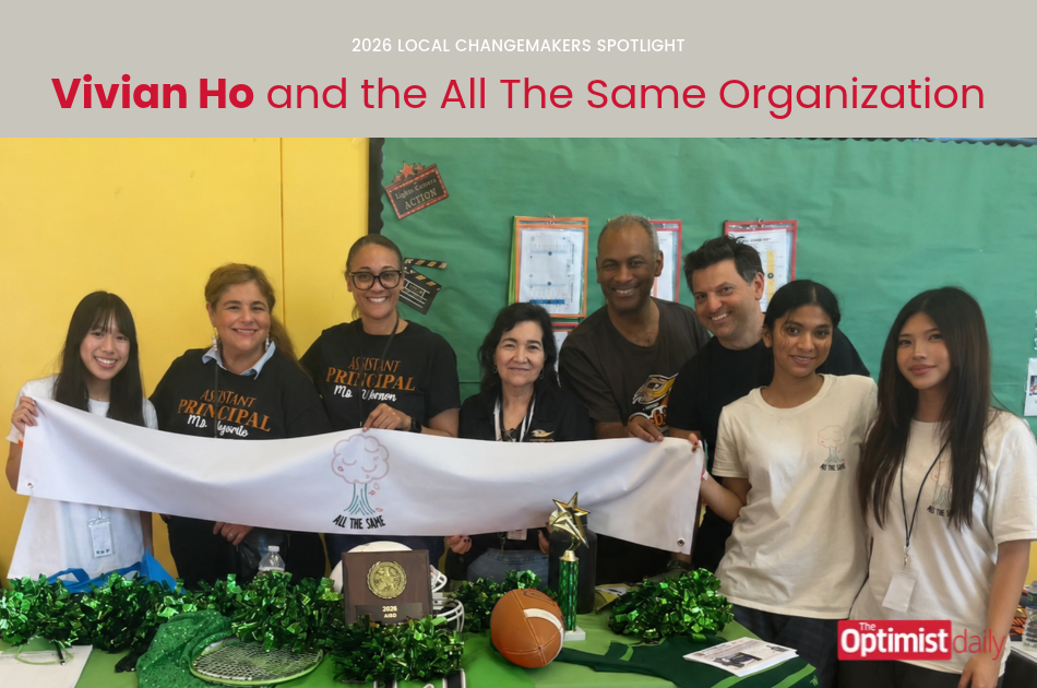 Group of eight diverse volunteers pose together, holding a white banner reading 'All The Same', with trophies, a basketball, and green decorations on a table at a community event.