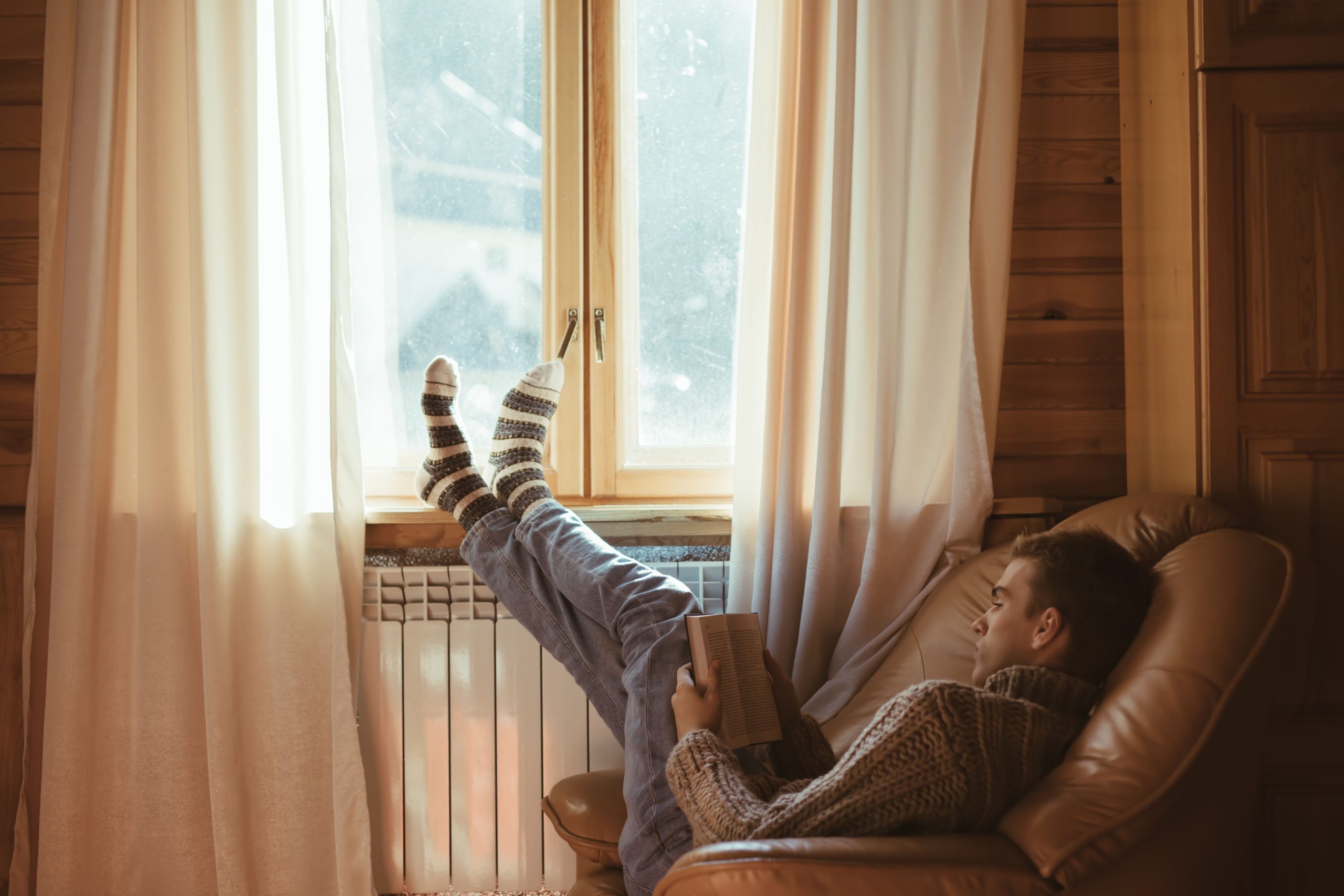 Young man in warm sweater reading book while relaxing on armchair by the window