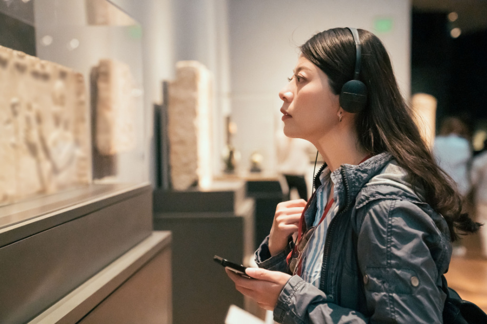 Young Asian woman looking at exposition in museum using headphones and listening audio guide in modern gallery. 