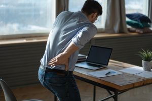Rear view young man suffering from sudden backache, getting out of uncomfortable chair at workplace, touching lower back, unhealthy businessman student office worker feeling discomfort