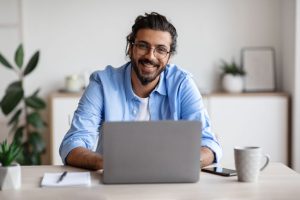 Successful Freelancer. Happy Millennial Indian Man Sitting At Desk With Laptop
