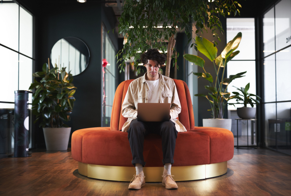 Full length shot of casually dressed young man working on laptop in modern open plan office sitting on orange circular sofa