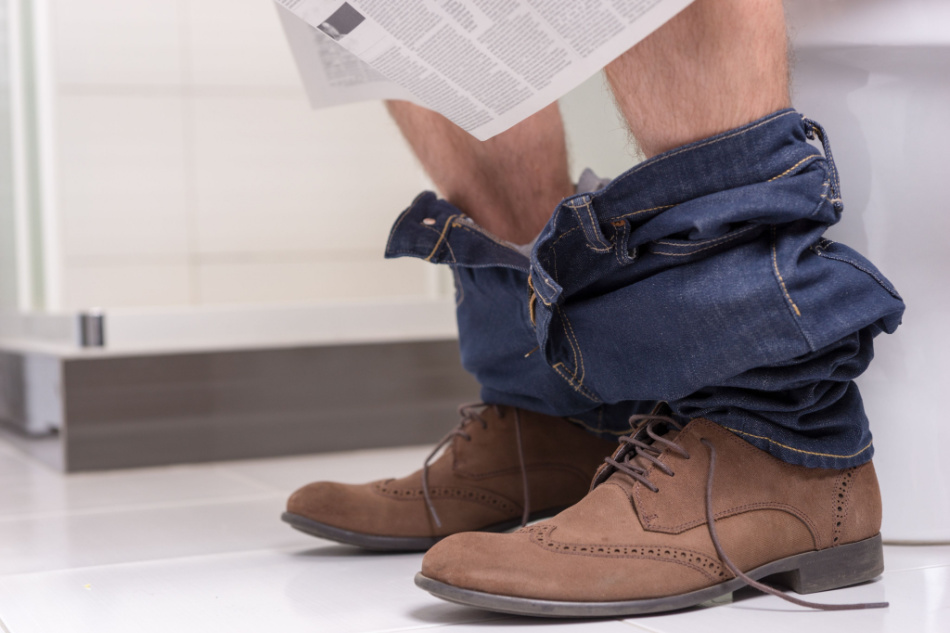 Close up view of man wearing jeans and shoes reading newspaper while sitting on the toilet seat in the modern tiled bathroom at home