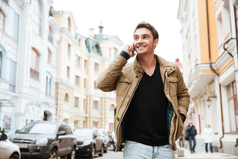 Image of happy young man walking on the street and looking aside while talking by his phone.