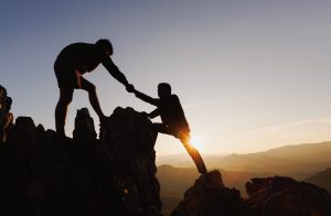 Two silhouetted hikers on jagged rocks helping each other reach the summit at sunset, mountain range in the distance