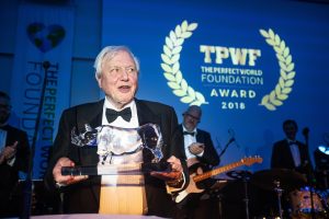 Older man in a tuxedo holding a crystal award at an awards ceremony; blue backdrop reads 'TPWF The Perfect World Foundation Award 2018'.
