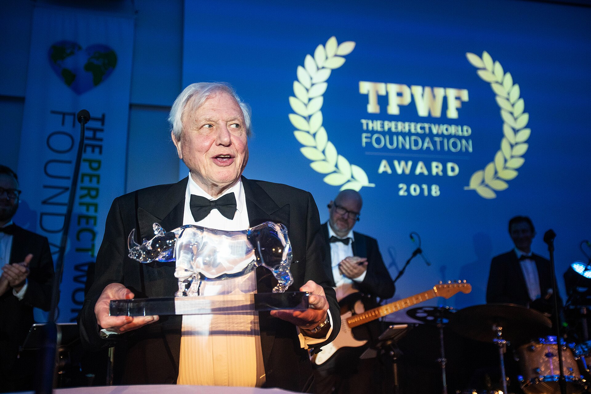 Older man in a tuxedo holding a crystal award at an awards ceremony; blue backdrop reads 'TPWF The Perfect World Foundation Award 2018'.