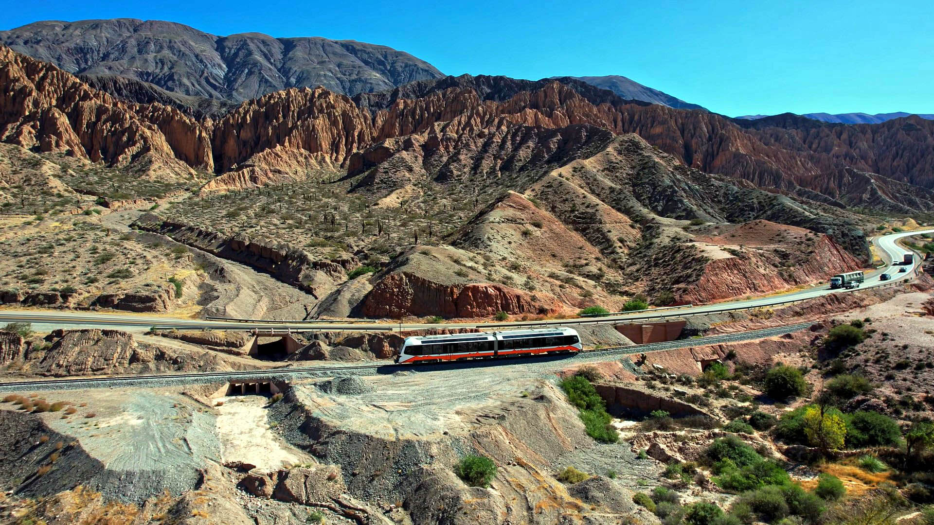Desert canyon with red layered rock, a white and red train on curved tracks in the foreground, and a winding road with vehicles on the right under a blue sky.