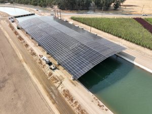 Aerial view of a large solar panel array over a canal, with farmland and a road nearby.