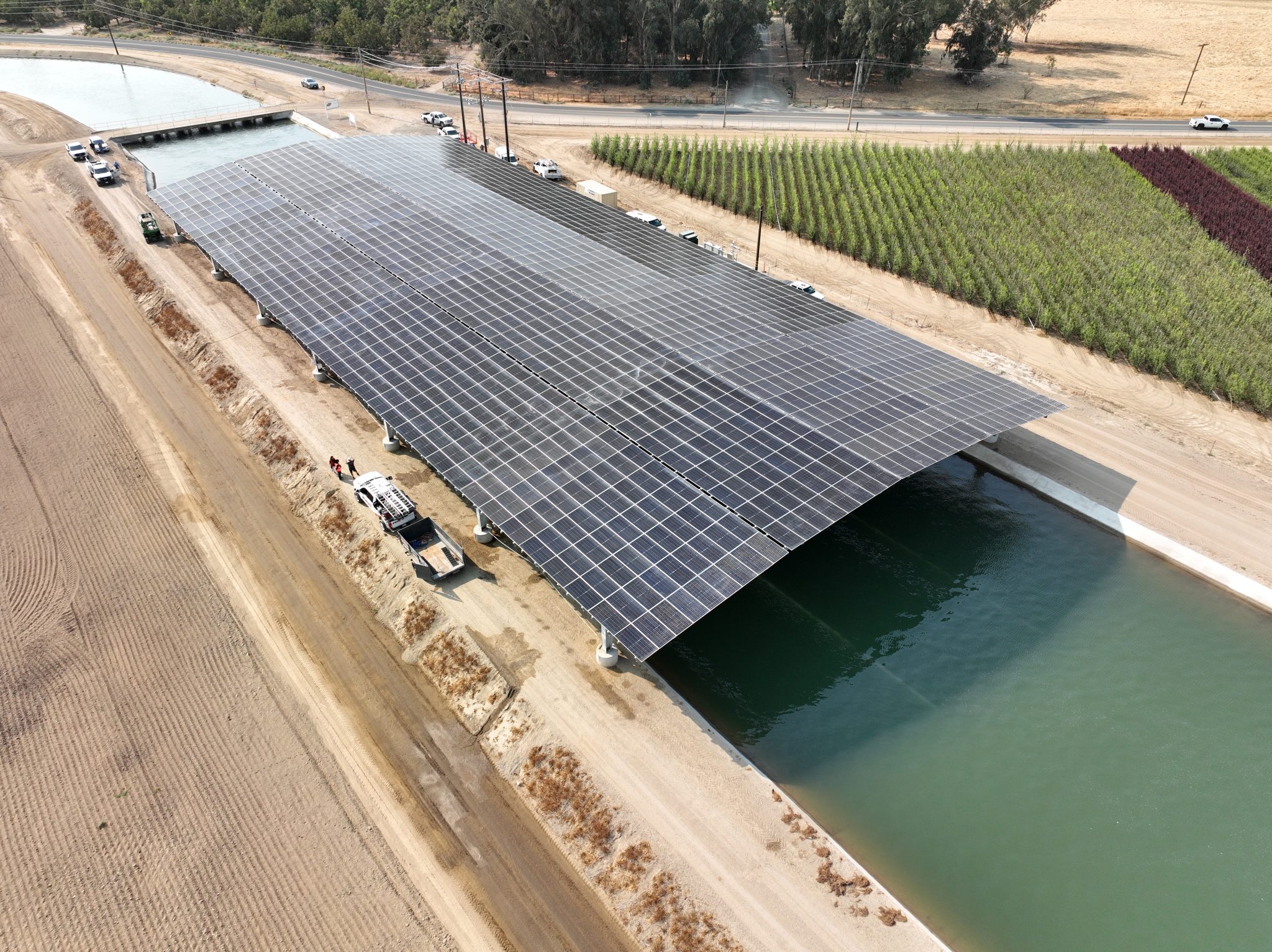 Aerial view of a large solar panel array over a canal, with farmland and a road nearby.