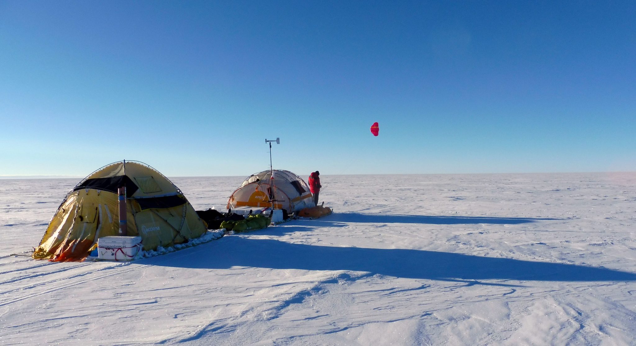 Polar camping scene: two tents on snow, gear scattered, a person in a red jacket near equipment, with a red paraglider overhead in blue sky.