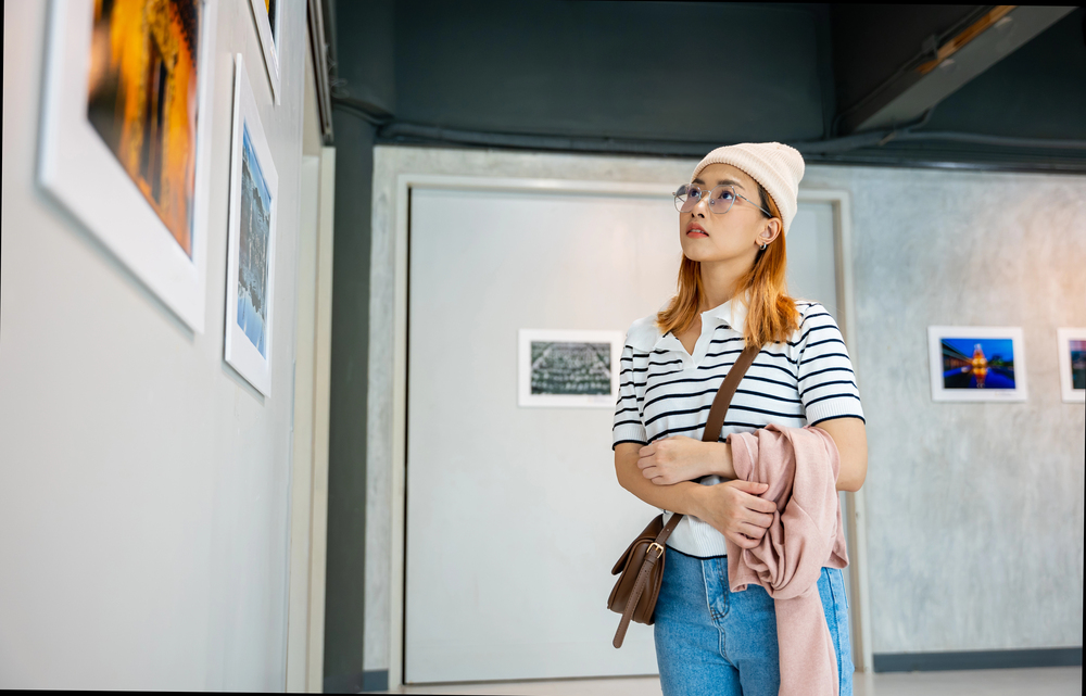 Woman in a striped shirt and beanie stands in an art gallery, looking at framed photos on the wall, holding a pink sweater and brown bag.