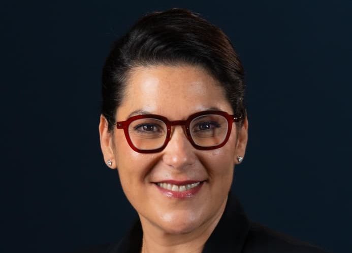 Smiling woman with dark hair, red round glasses, pearl earrings, wearing a black blazer against a dark blue background.