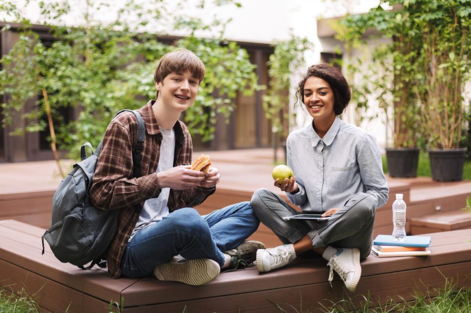 Couple of smiling students sitting on bench with sandwich and green apple and happily looking in camera while spending time together