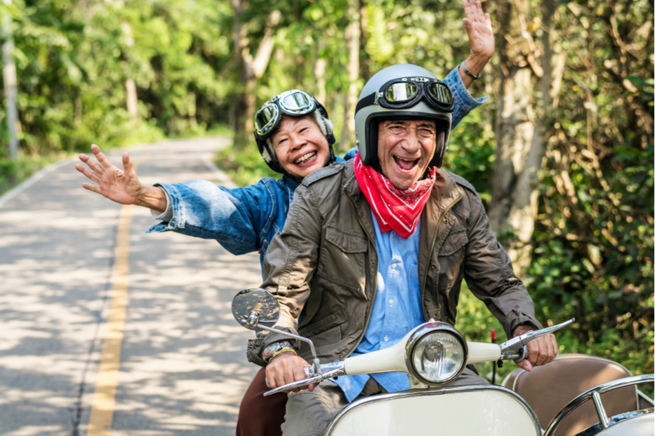 two older people happily driving on a road flanked by greenery on a moped