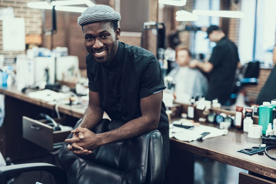 attractive and young Black barber master looking at camera standing against blurred barbershop
