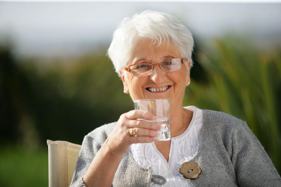 Elderly smiling white woman sits outside and raises a glass of water.
