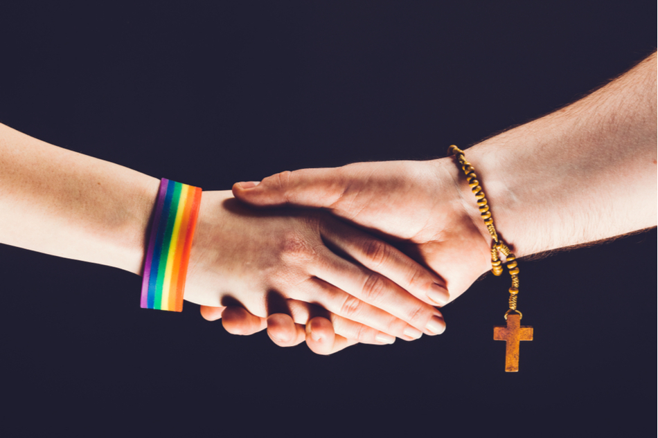 two people, one wearing LGBTQ colors and the other with a crucifix, shake hands