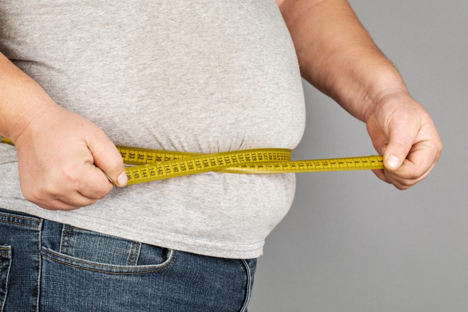 A person in a grey t-shirt measures their belly fat with a yellow measuring tape.