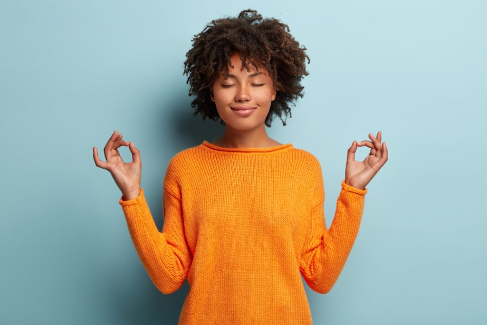 Mindful peaceful woman meditates indoors while keeping hands in mudra gesture, has eyes closed, and is wearing orange top in front of a blue background.