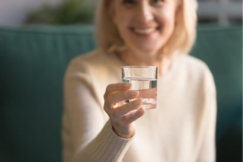 woman stretch arm to camera holds glass of water close up focus on beverage and arm, healthy lifestyle good habits