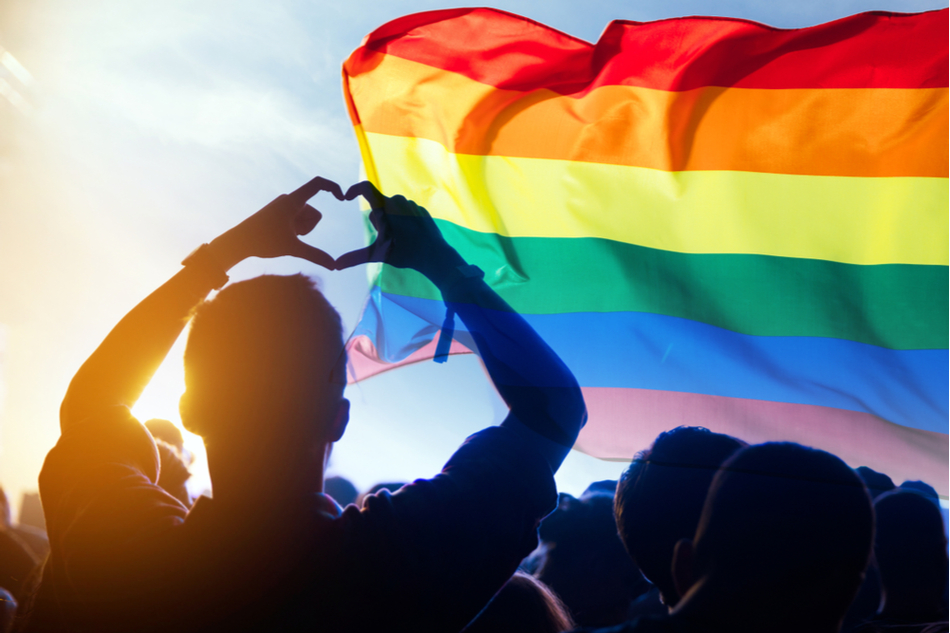 LGBTQ rainbow flag flies against the sky, silhouette of a person making a heart with both their hands
