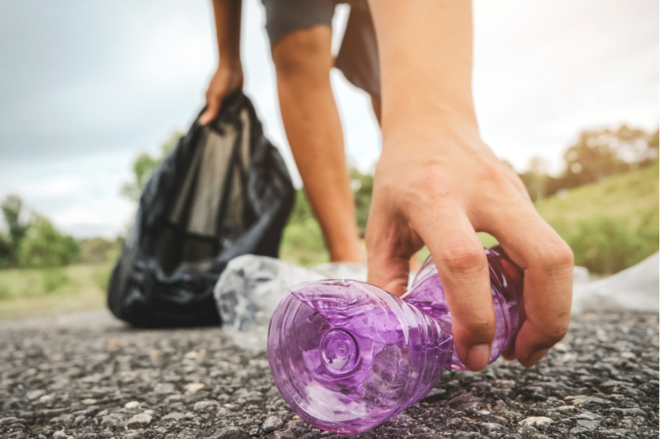 man's hand picks up littered purple plastic bottle from roadside