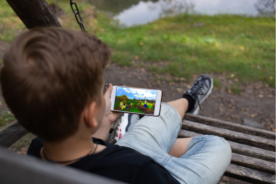 The boy sits on a bench and plays the game online Minecraft in the Park