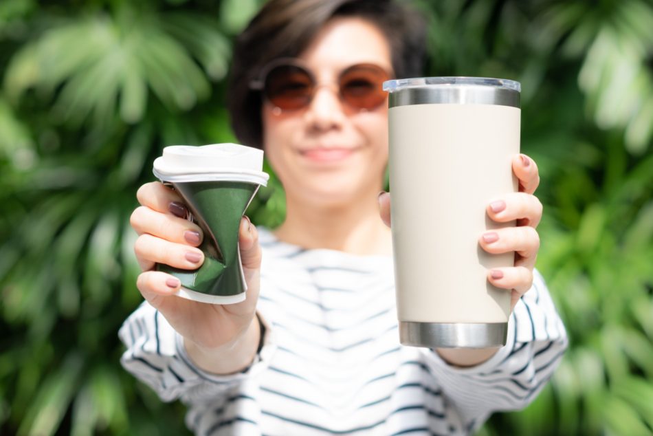 Person holding takeaway coffee cup in both hands, one is a single use paper cup with plastic lid the other one is a reusable stainless tumbler.