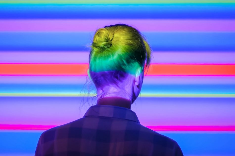 Back view of young woman looking around at modern immersive exhibition or club event with changing multi color projector light