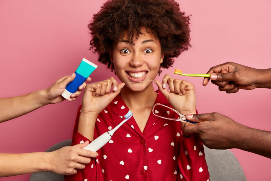 Shot of curly woman cares about teeth, holds dental floss, surrounded by toothpaste and toothbrushes.