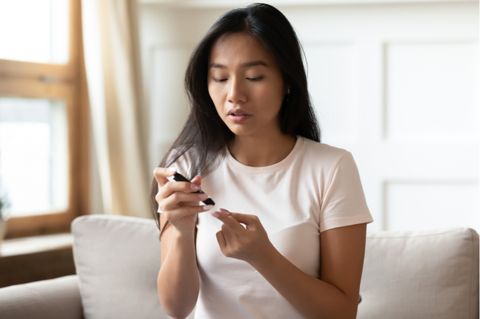 Young diabetic Asian woman sits on couch and pricks finger to measure blood sugar level