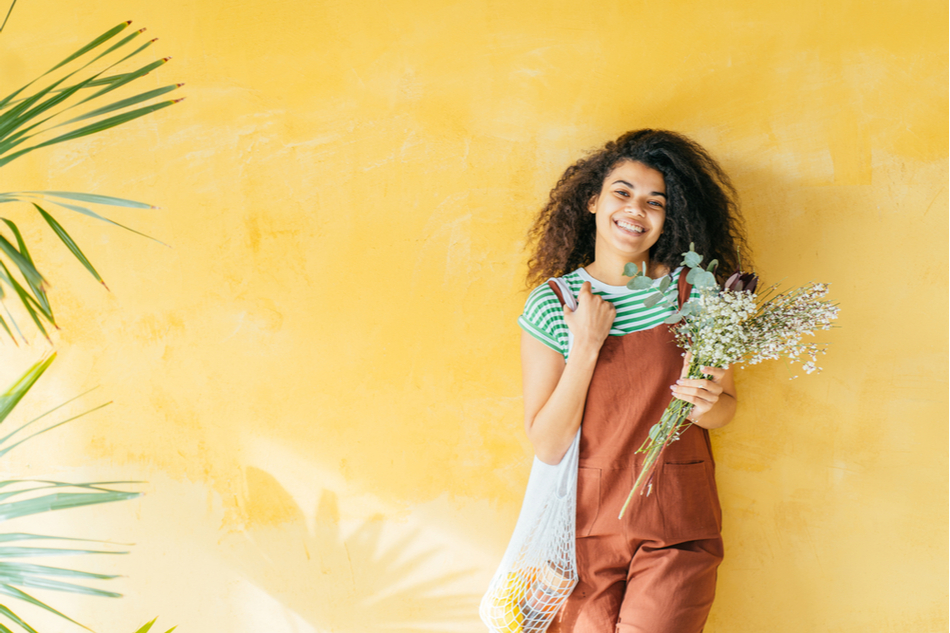 smiling mixed-race woman stands smiling against a yellow backdrop with a reusable bag full of plant-based groceries