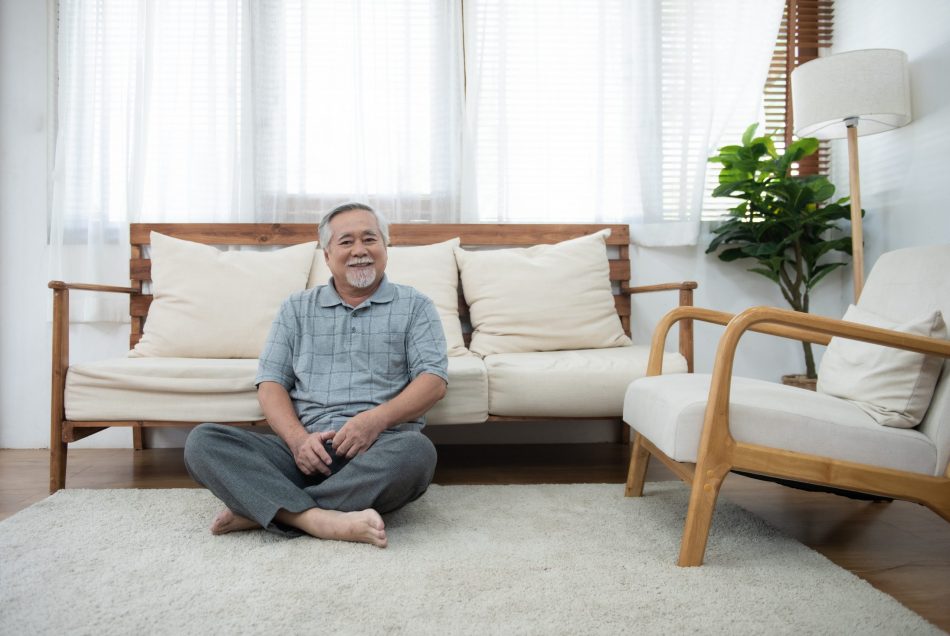 elderly white man sits cross legged on the floor in living room