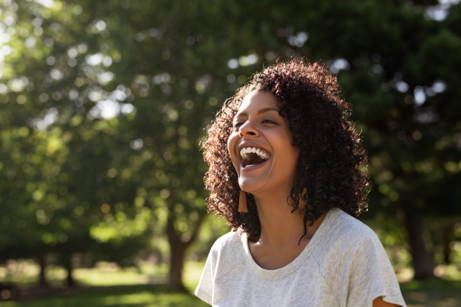 Young woman with curly hair laughing while standing outside in a park on a sunny summer afternoon.