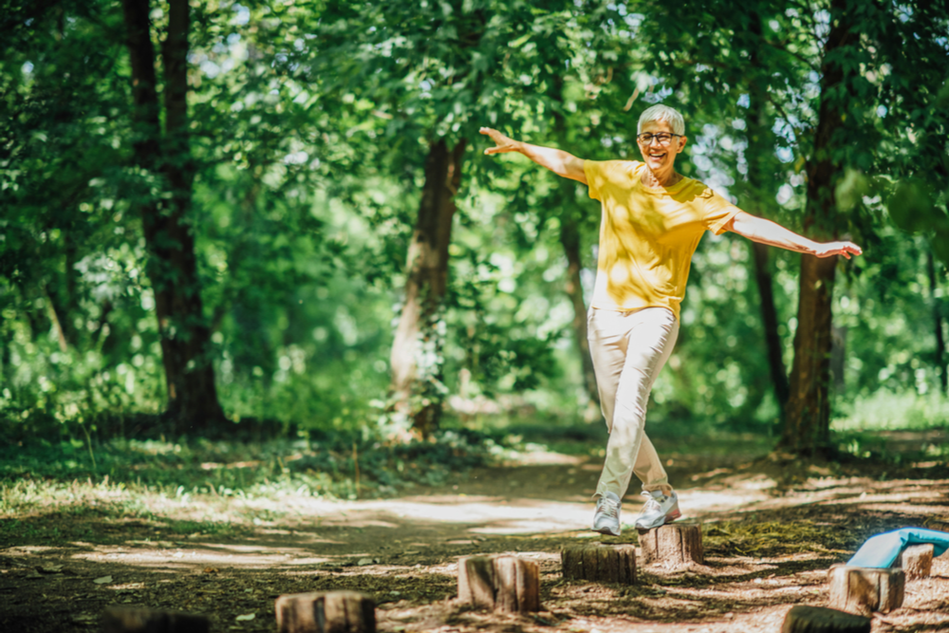 older woman in yellow shirt balances on tree stumps in a forest