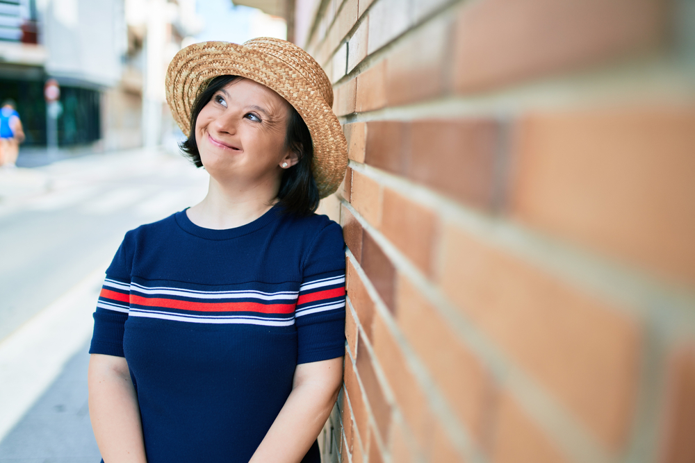 Brunette woman with Down syndrome leans against brick wall in a sunhat
