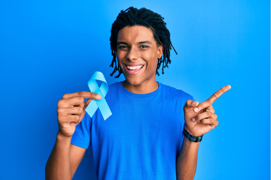 young happy Black man in blue shirt against blue background holds blue prostate cancer ribbon